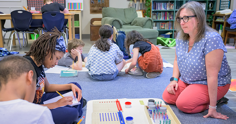 Montessori teacher works with elementary-age students during a classroom learning activity, as students write, collaborate and use Montessori materials on the classroom floor.
