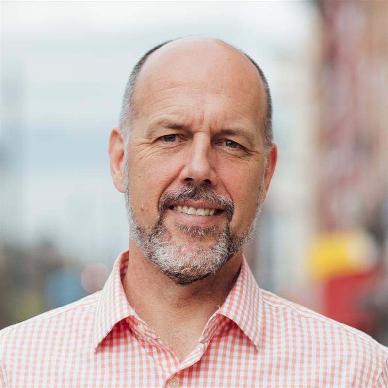 Headshot portrait of Stephen Metraux smiling and standing outdoors while wearing an orange and white checkered dress shirt, a blurred city block environment in the background