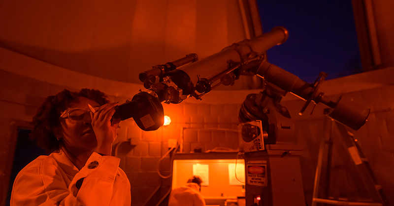 A student inside an observatory looks through a telescope pointed at the night sky.