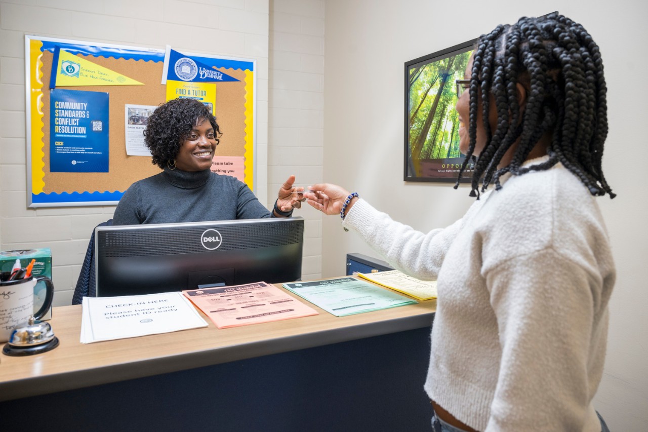 A college student presents her ID card to a staff person who stands behind a counter of the College of Arts & Sciences Student Success Center and uses a computer to check her in.