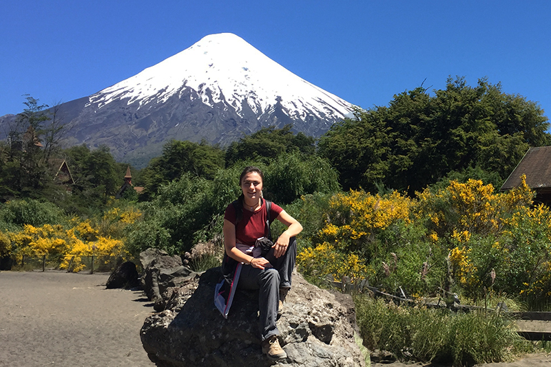 University of Delaware dressed in hiking clothes sits on a rock on a sunny day in Chile with a large, snow-capped volcano in the background.