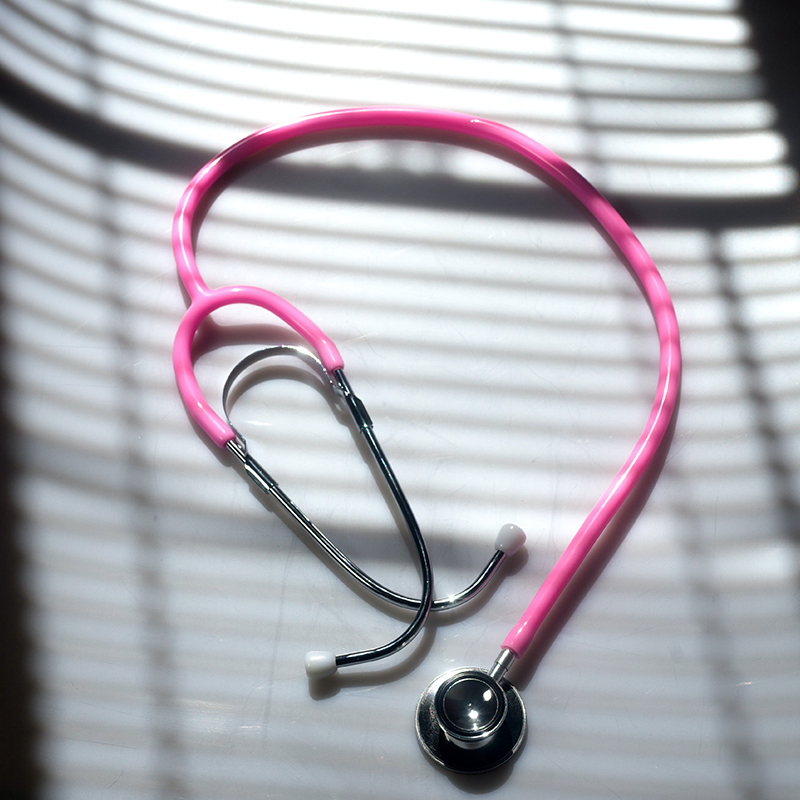 Doctor's pink stethoscope rests on a white surface, covered by a shadow of window blinds.