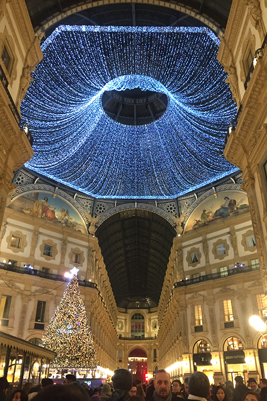 The Galleria Vitorio Emanuele in Milano, Italy, decorated at Christmas with blue lights at the top of the atrium which is filled with historic European style buildings with frescoes and a crowd of people walking around.