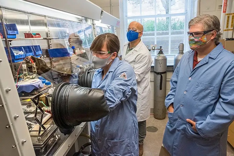 Graduate student Amanda Weaver setting an electrochemical process in motion in a lab setting. Two professors are behind her observing.