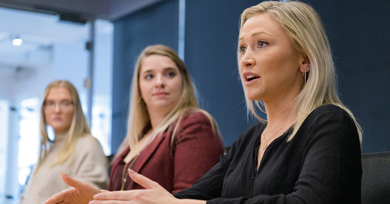 A woman speaks persuasively during a meeting inside a conference room while two other women listen attentively.