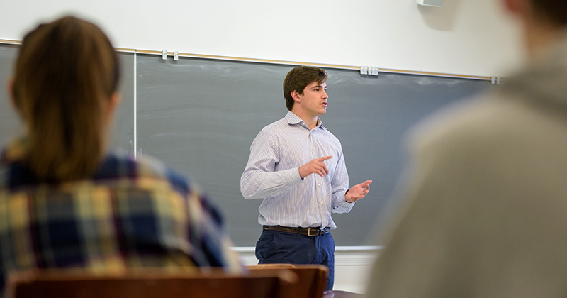 A graduate student dressed in business attire stand in the front of a classroom and presents a lecture to undergraduate students.
