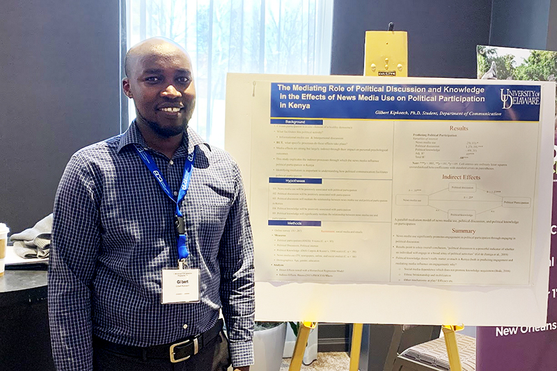 A college graduate student stand inside an office next to a research poster on an easel.