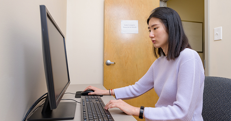 A female graduate students sit inside a small office and types on a computer keyboard in front of a desktop computer.