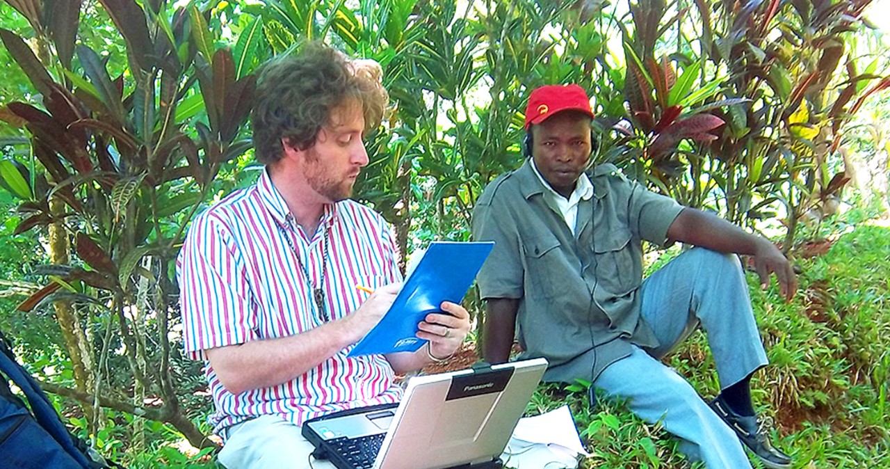 A college student with a laptop and backpack next to him sits under some trees and interviews a man wearing a red baseball cap.