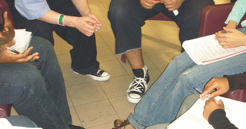 Group of people are sitting and facing each other in a circle, we only see their legs and feet, they are weaing jeans and sneakers