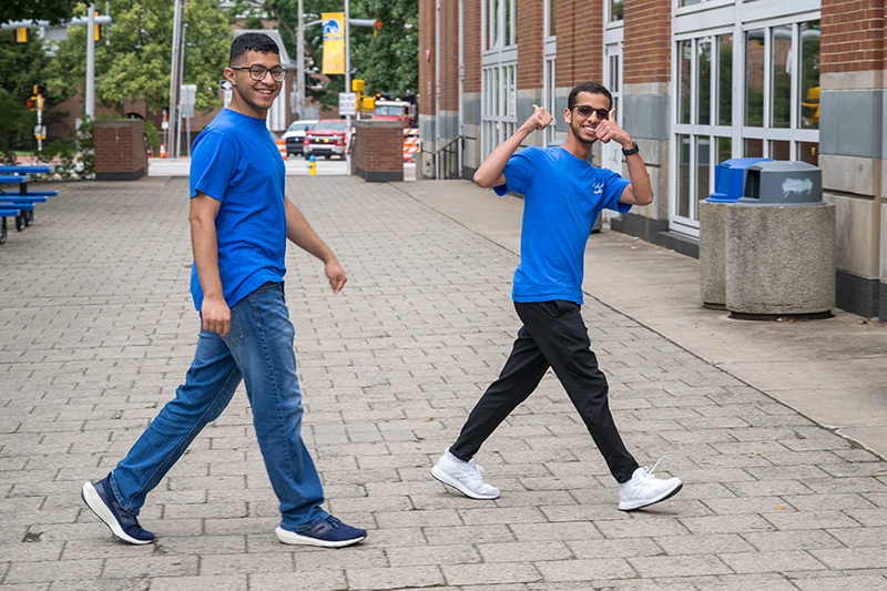 Two college students wearing matching blue shirts walk on a sidewalk on a college campus.
