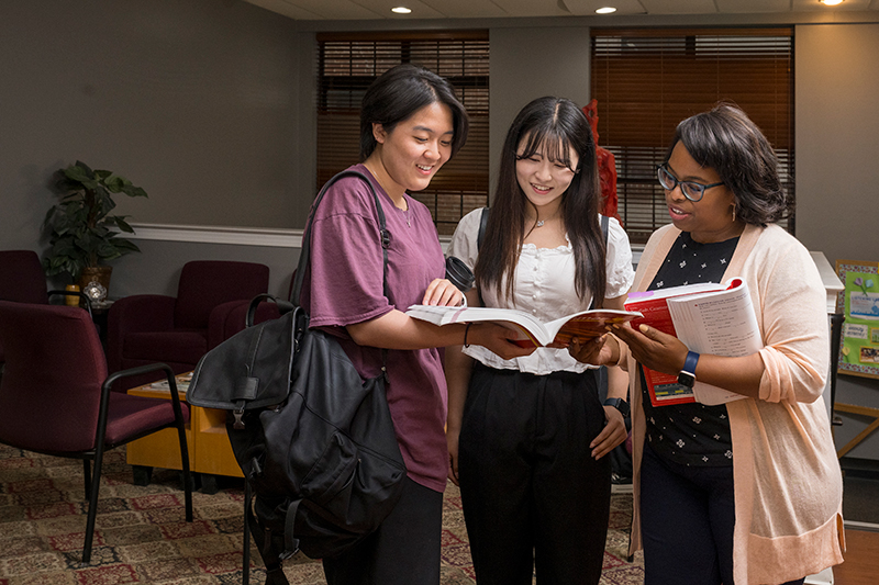 Two college students and a professor stand inside a meeting room and review a book together.