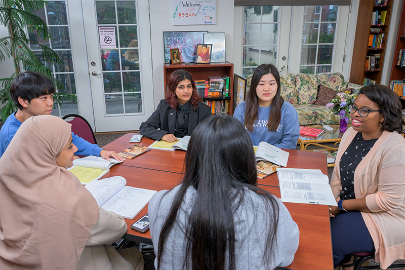 Five college students and their instructor discuss their coursework while sitting around a table with their books and papers.