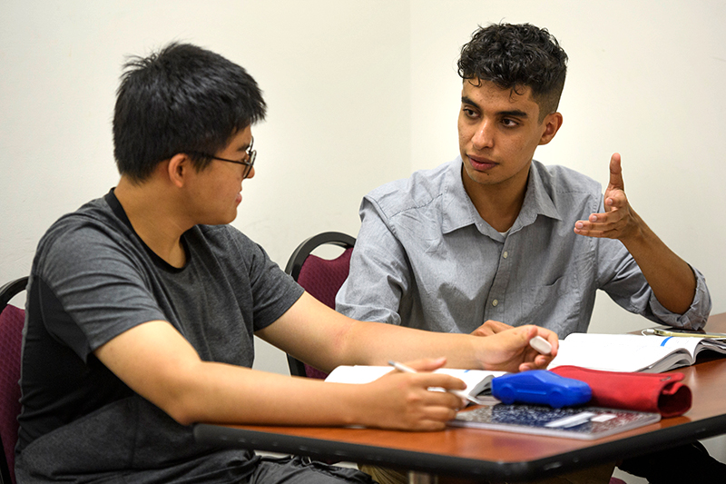 Two college students talk to each other while sitting at a desk with notebooks and papers.