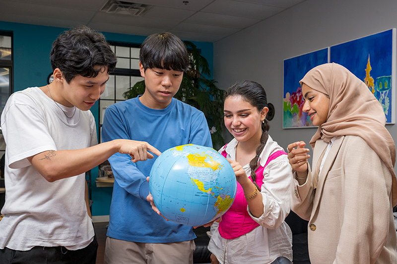 A diverse group of four college students hold an inflatable world globe and point to different areas on the map.