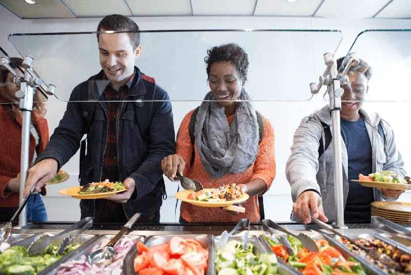 Three college students build their own salads at a salad bar in a cafeteria.
