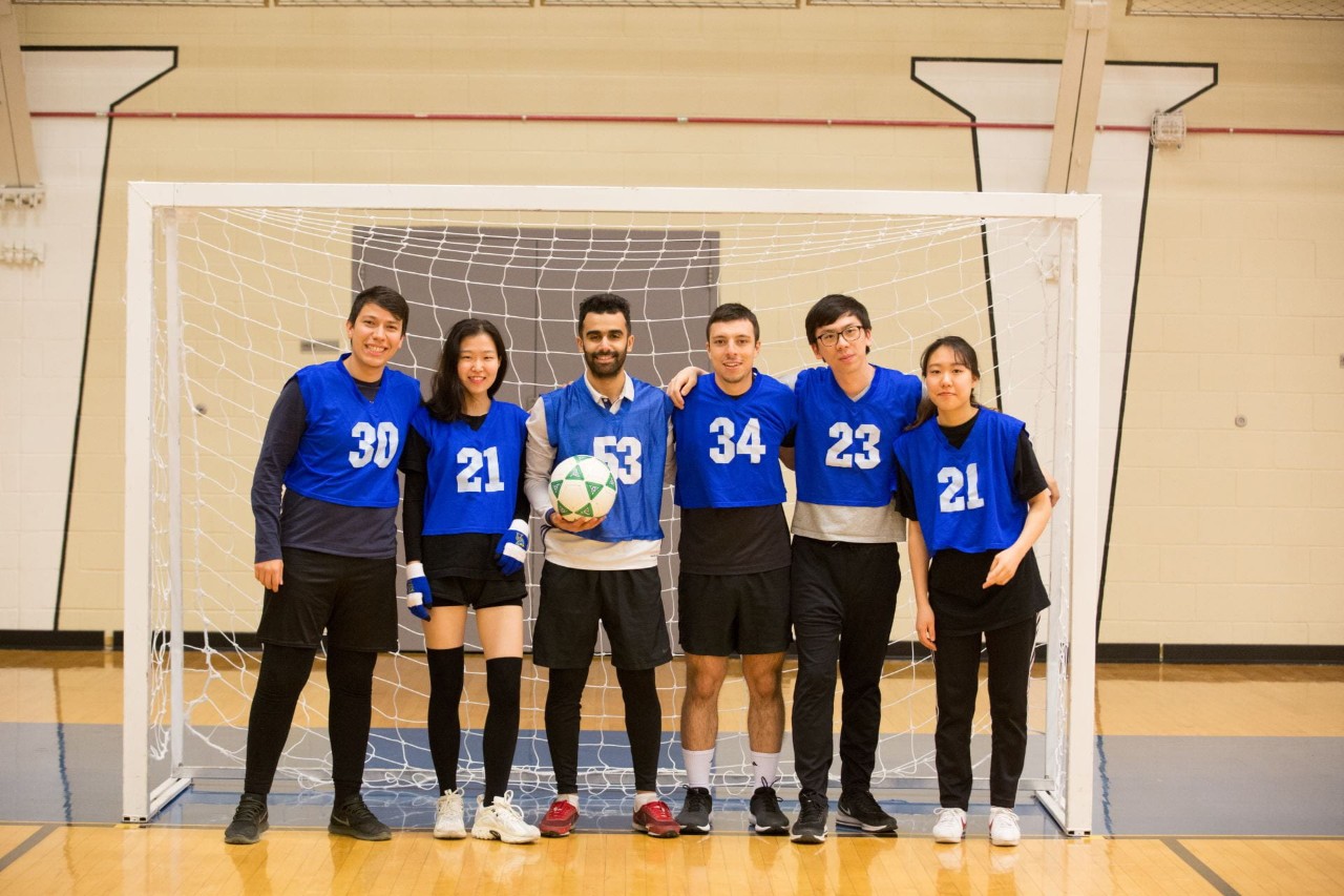 Six international students in blue and black uniforms standing in front of a goal with a soccer ball