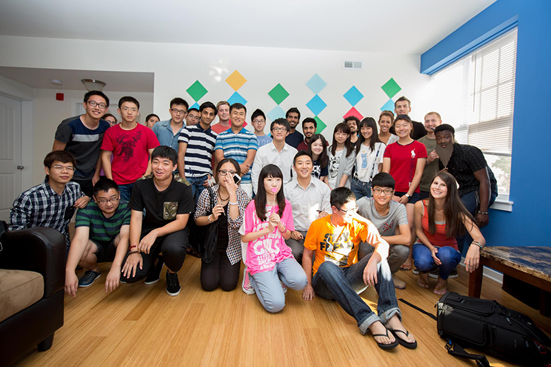 A large group of smiling college students pose together for a group photo inside an apartment.