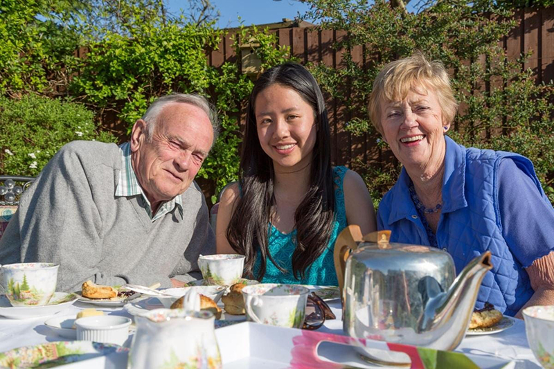 A smiling college student sits between her homestay hosts at a table outside during a tea party.