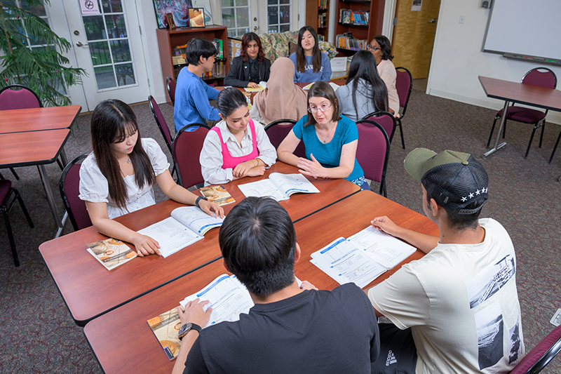 Four college students and their instructor sit inside a classroom at a table with their books and discuss their coursework.