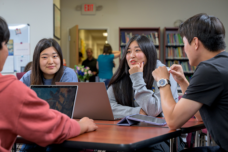 Four college students talk to each other around a table inside a classroom.