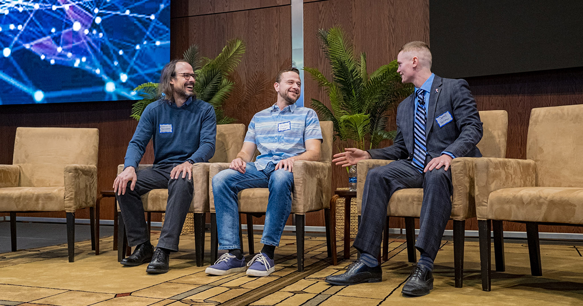 Three male presenters sitting on a stage in discussion.