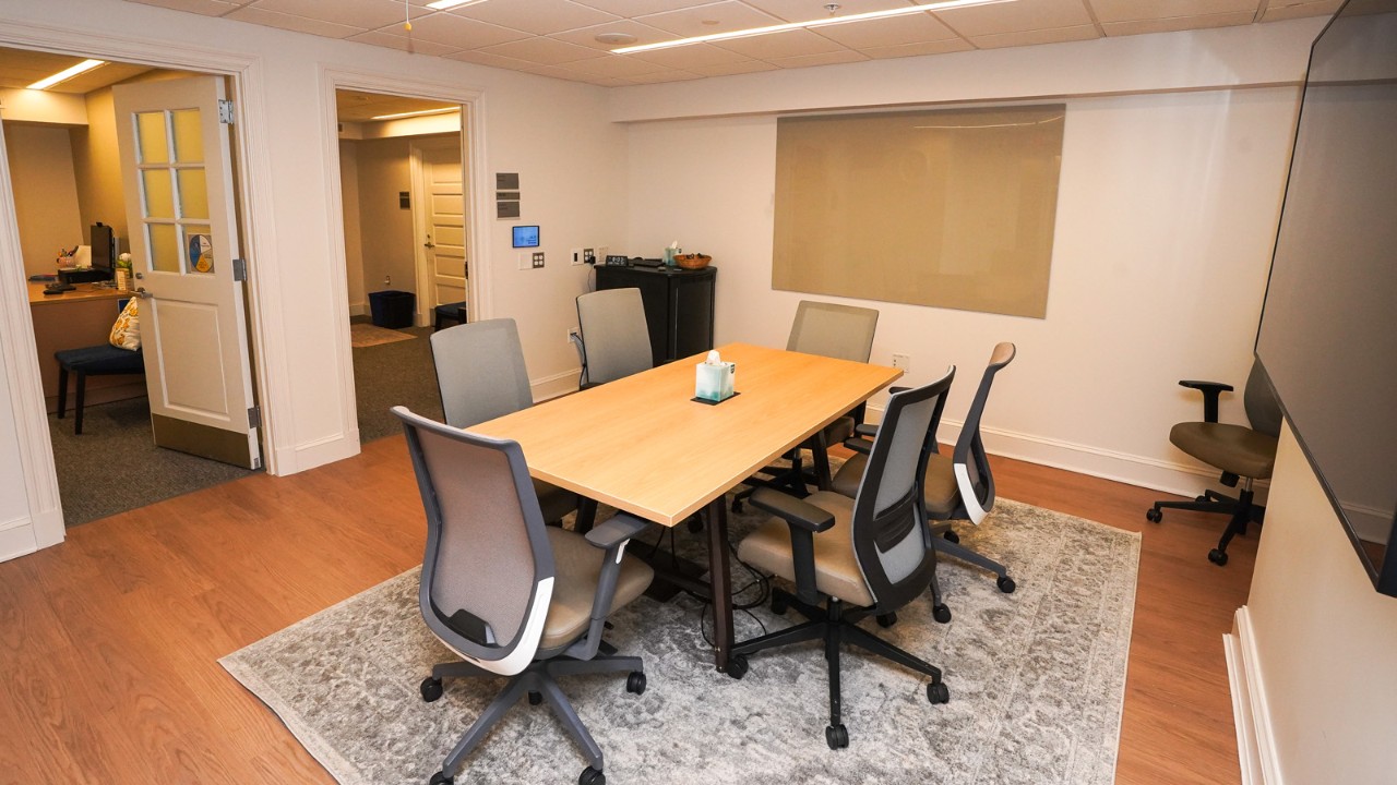 a conference table sits on a rug in a room with a monitor mounted on the wall