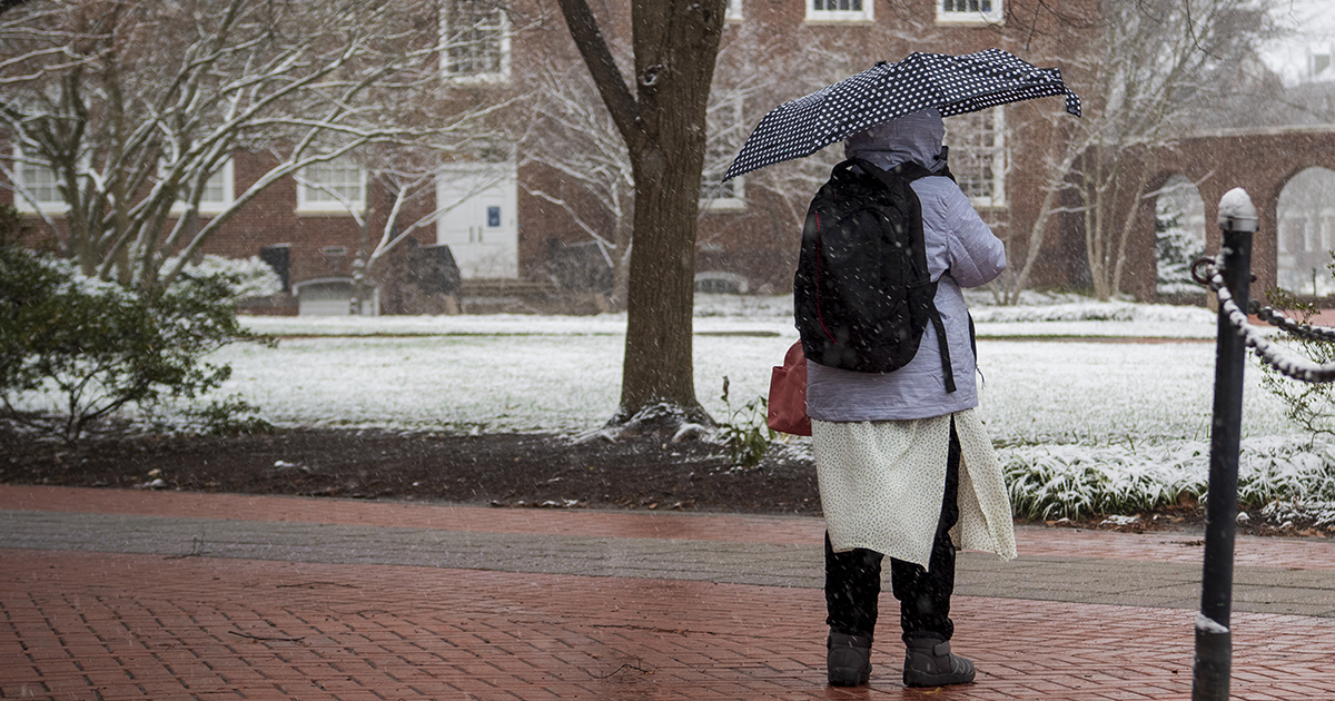 Student in puffy coat with backpack walks across snowy campus holding umbrella