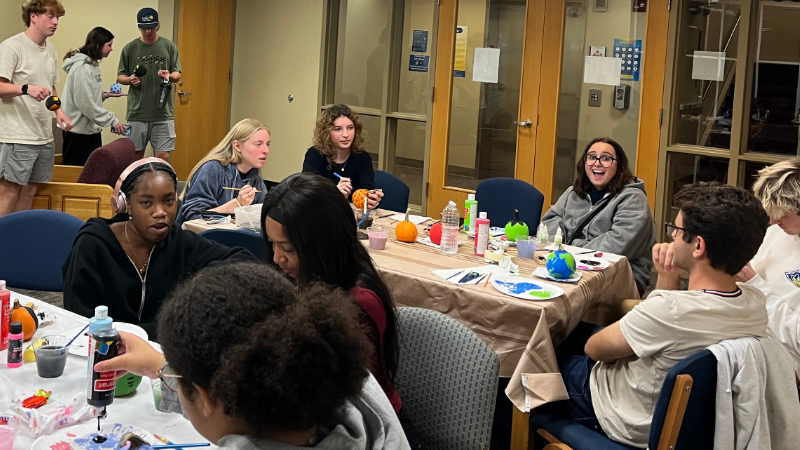 Students sit around tables in a residence hall lounge painting pumpkins