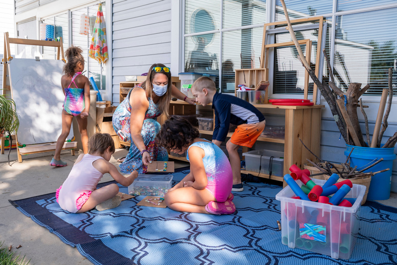 The new procedures to the Early Learning Center (ELC) include temperature checks at drop off, no parents allowed in the buildings and new outdoor classroom learning spaces. Pre-K teacher Cindy Hartsoe made her own outdoor space while the ELC was shut down.  Here she is with students: Marshall-navy shirt, Kylie-rainbow swimsuit, Ila-princess swimsuit, Sofia-pink.