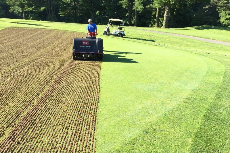 An employee operates a turf aerator at Deerfield Golf Club in Newark, Delaware. Aeration keeps the playing surface healthy by removing tiny soil cores. These holes enable moisture evaporation and promote gas exchange in the soil. The process results in stronger root systems.